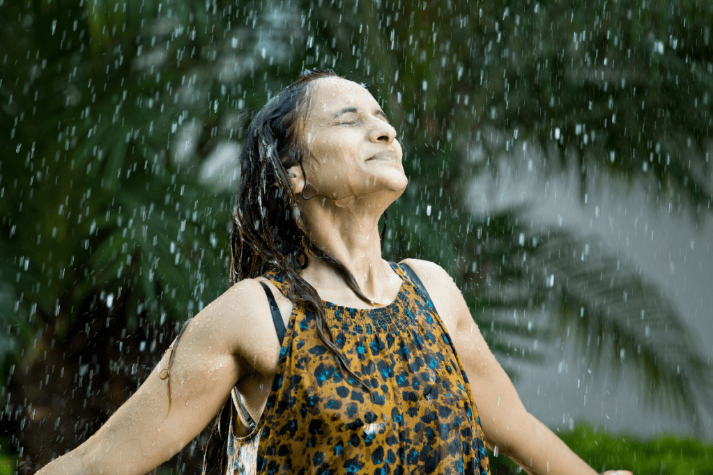 A woman stands in the rain with her eyes closed. Her dark hair and clothes are wet.