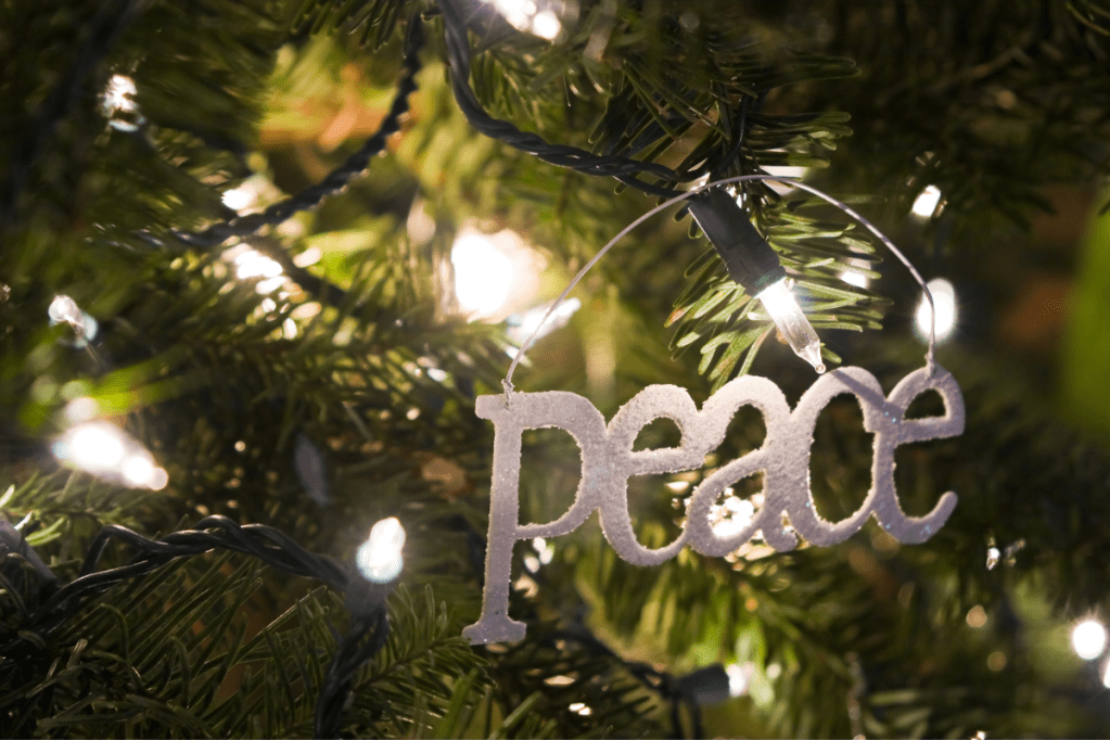 A closeup of a christmas tree, featuring fairy lights and an ornament that reads 'Peace'