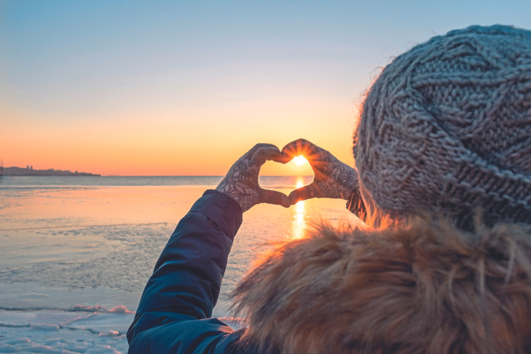 A woman at the beach holding her hands up in a heart, to frame the sunset over the sea