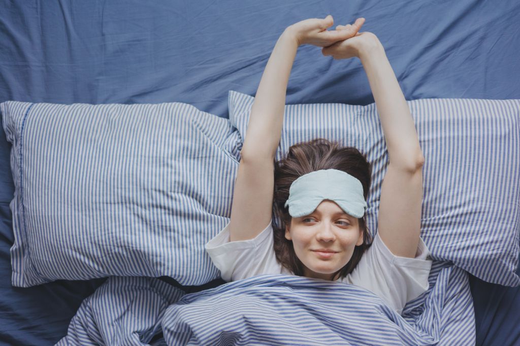 woman with face mask on stretches arms in bed