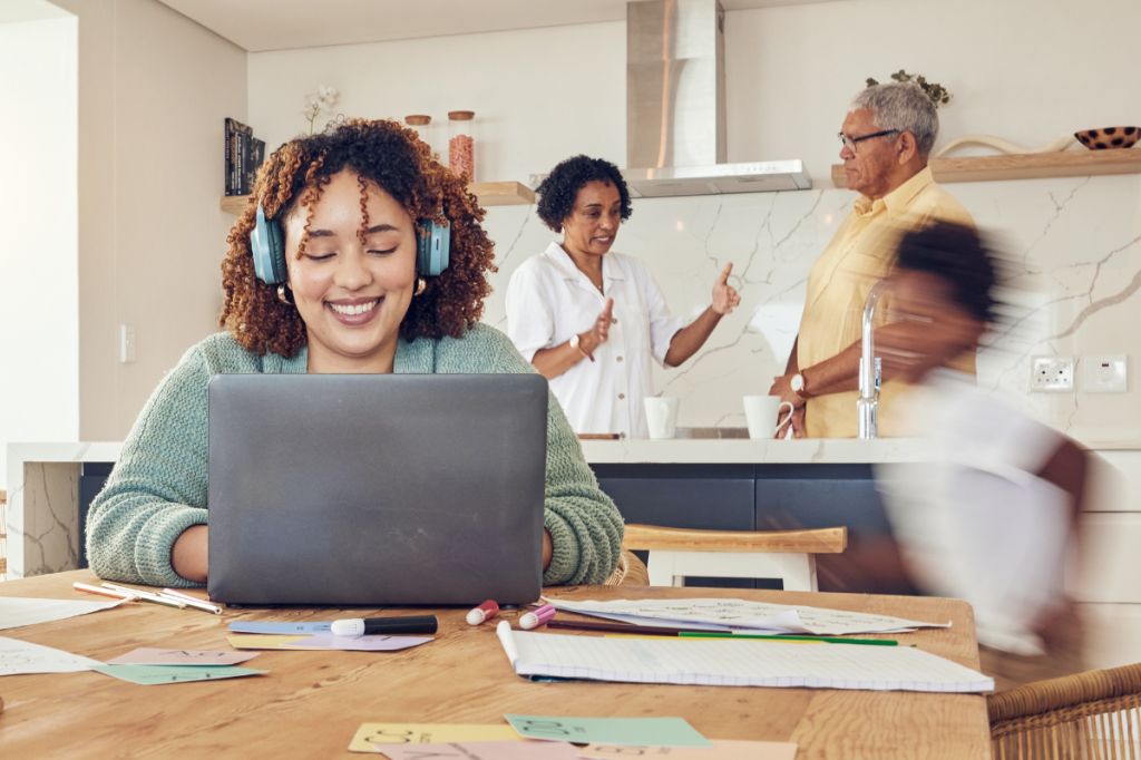 Mum is on computer with headphones on while people talk in background and child rushes by