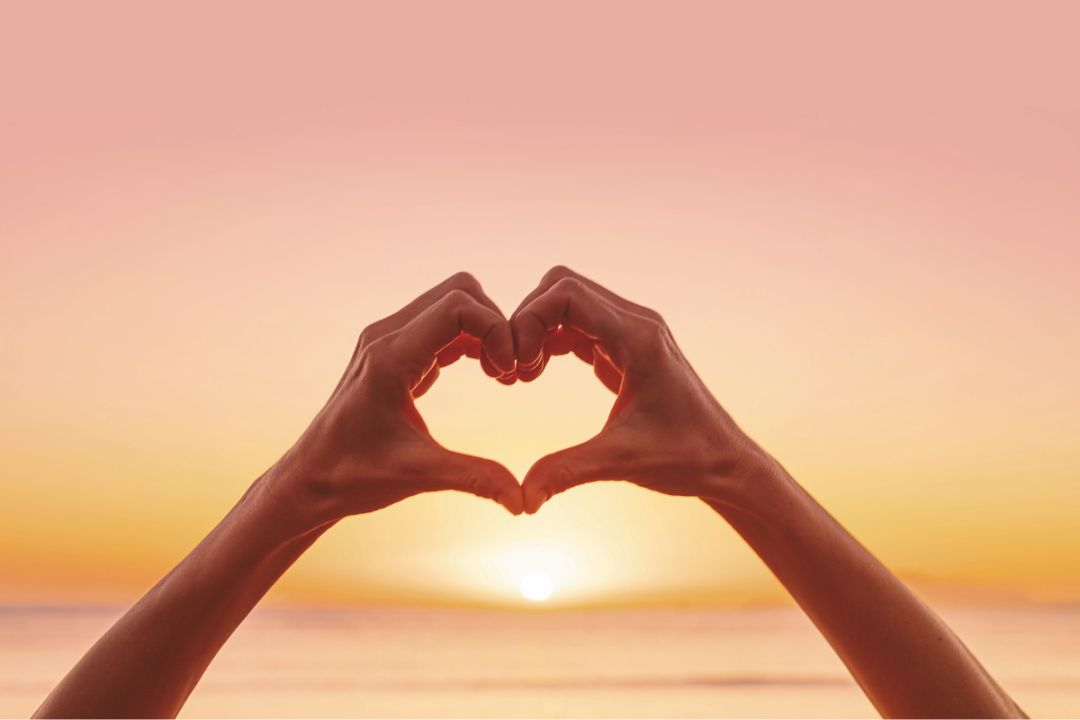 Hands make heart sign in front of beautiful sunset, symbolising kindness