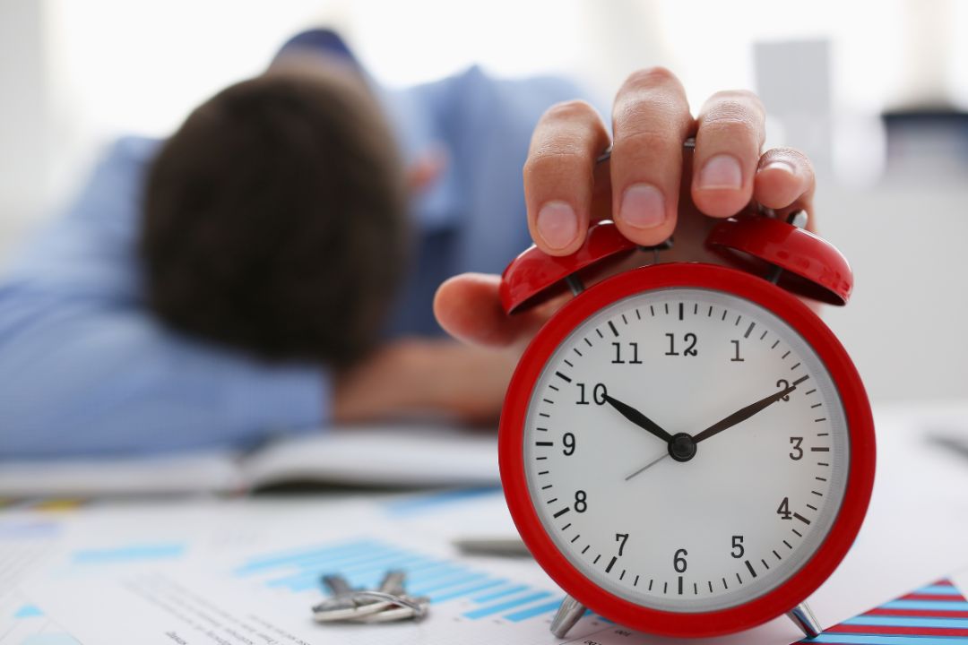 man has head on arms and hand on alarm clock