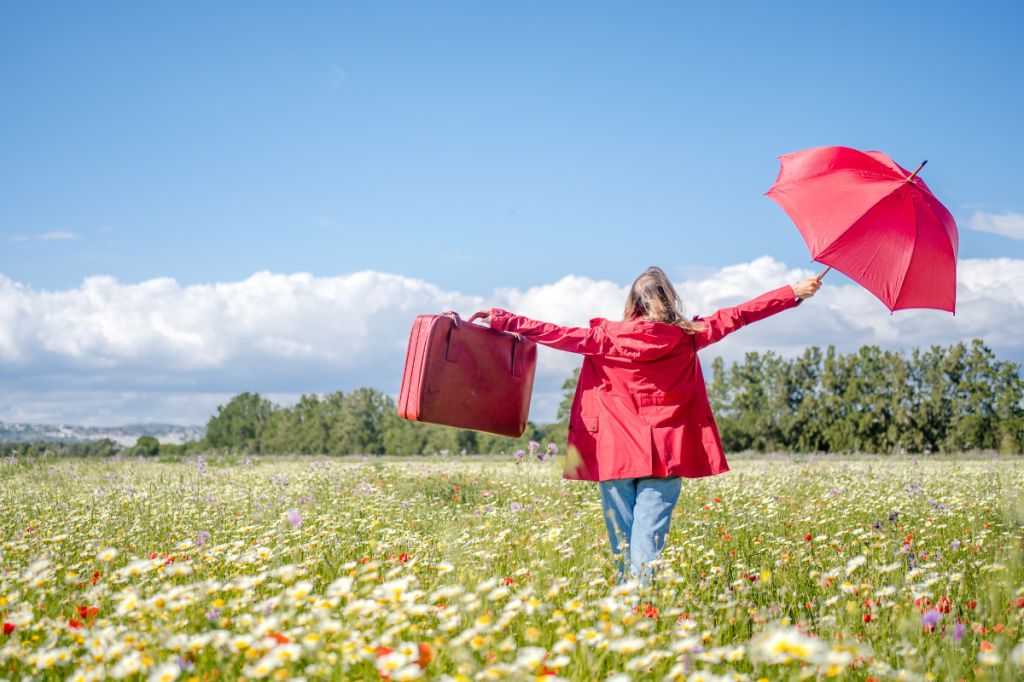 Woman in red coat walks through field of wildflowers carrying red umbrella and suitcase