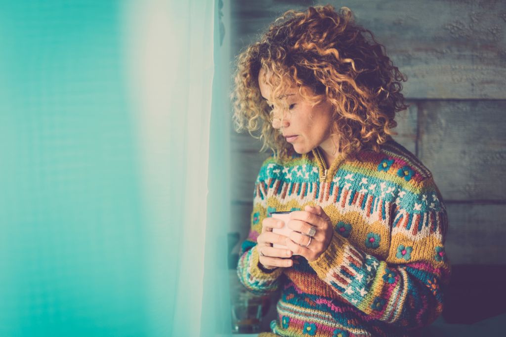 A woman with curly light brown hair holds a mug and looks down thoughtfully. She is wearing a multicoloured and patterned knitted cardigan that is zipped up.