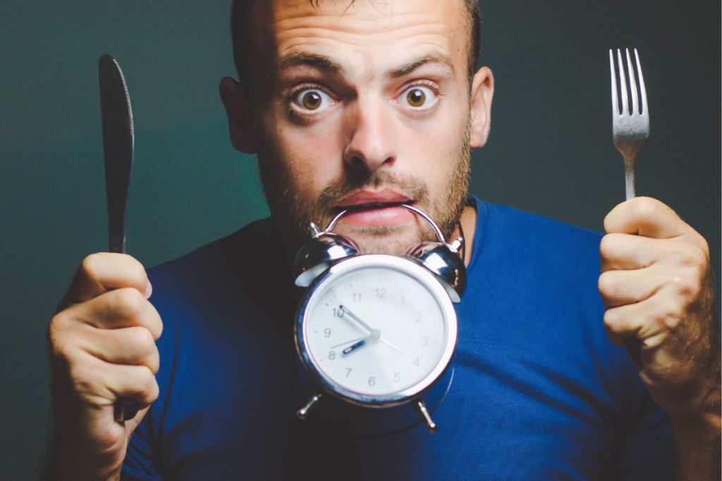 Man looks stressed, with alarm clock hanging from his teeth and knife and fork in his hands
