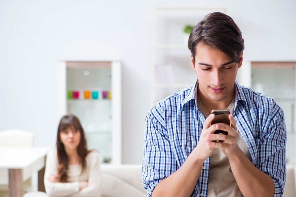 man sat looking at phone while girlfriend sits crossed arms in background