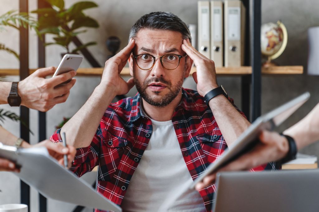 Man looks overwhelmed being handed computers, phones and tablets