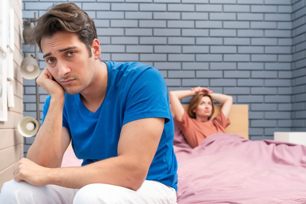 man sits on bed looking sadly into camera, frustrated girlfriend in background