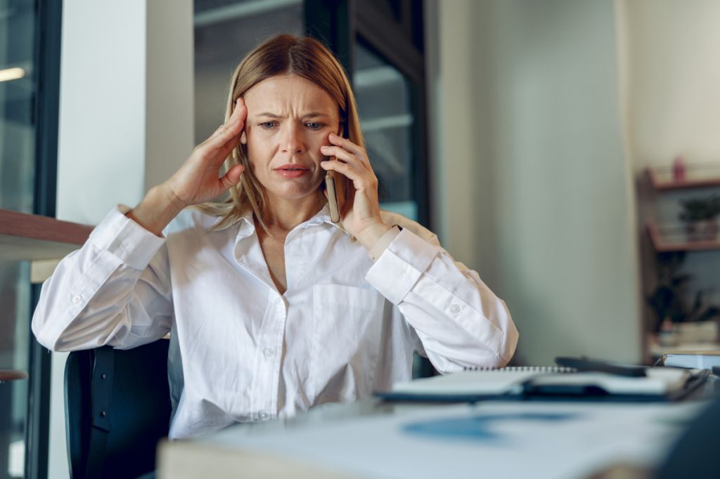 woman sits at desk, talks on phone looking very stressed