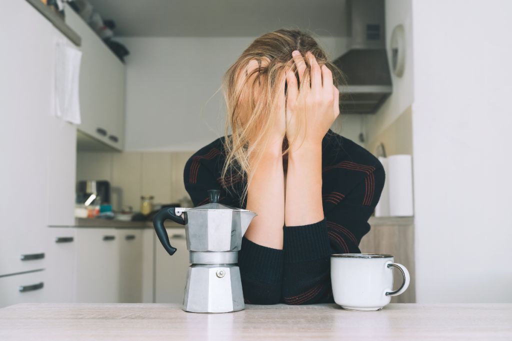 women covers face with hands with elbows on kitchen counter in front of coffee pot and mug