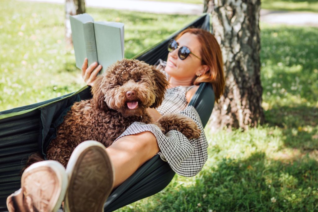 woman relaxes in a hammock, reading a book and cuddling a dog