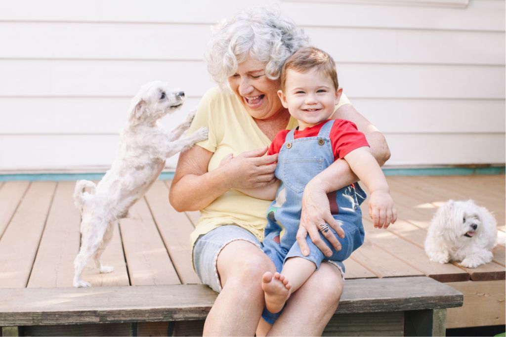 grandma cuddles grandson while cute white pup jumps up to greet her