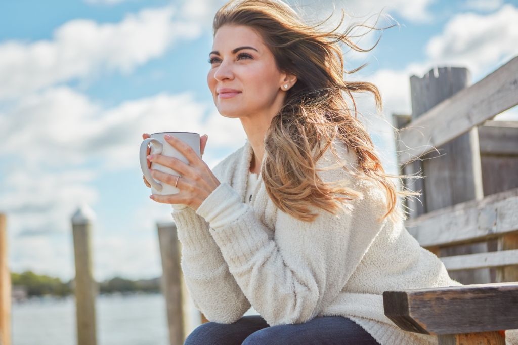 woman leans on fence and holds cup of coffee while smiling and enjoying the view