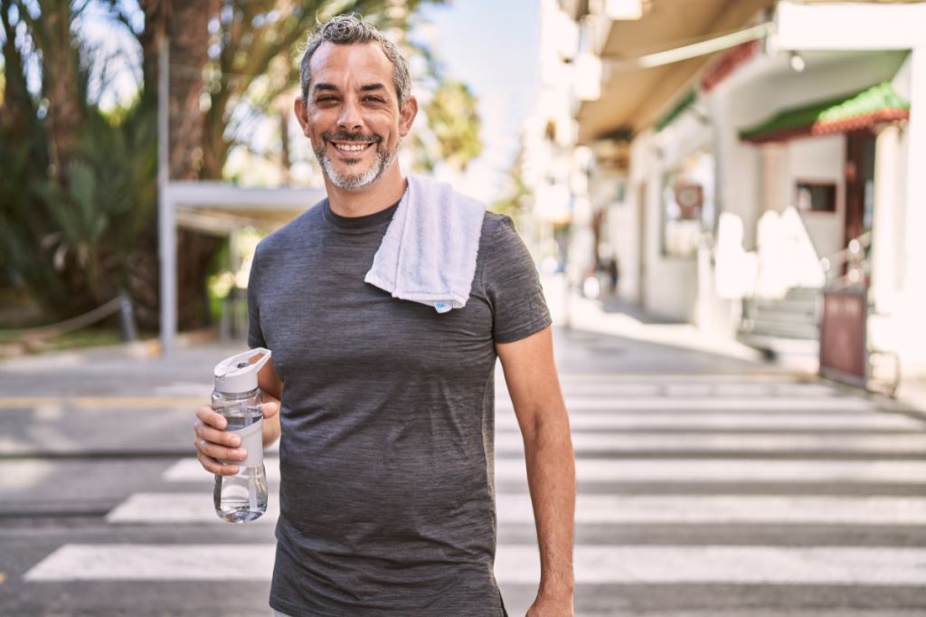 man sands on street with bottle of water smiling, looks like he has just completed a run