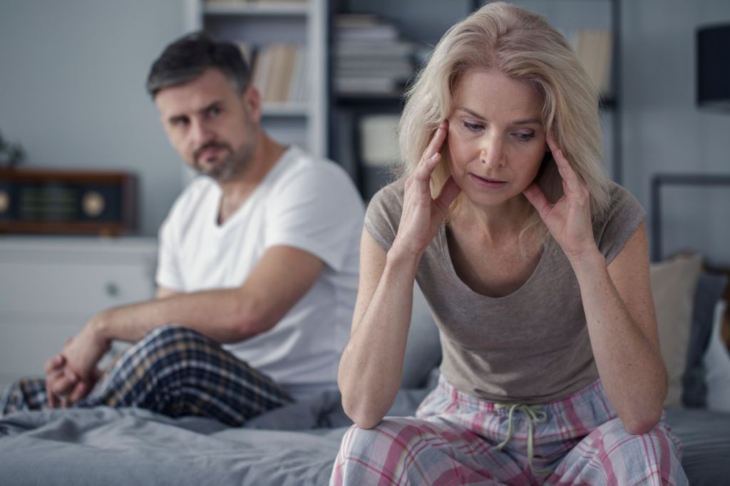 Couple sit on bed, woman can't look at man, he looks aggressively towards her