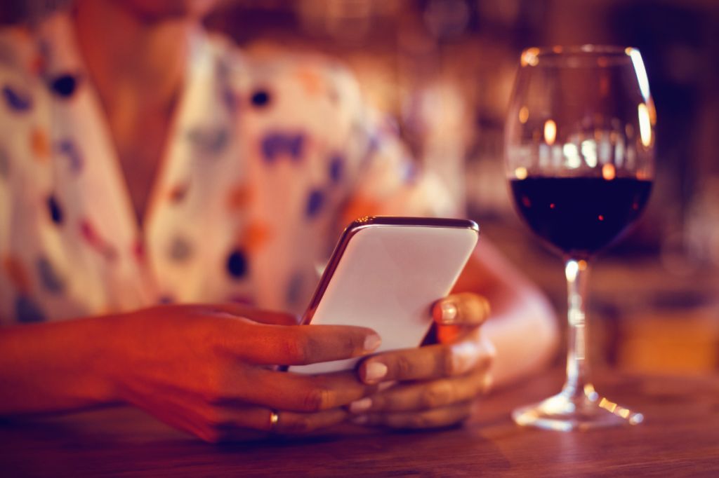 woman sits in bar with glass of wine by her absorbed in her phone