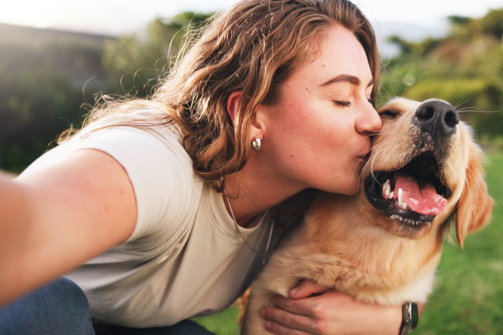 woman looks overjoyed to be kissing a golden retriever, selfie-style