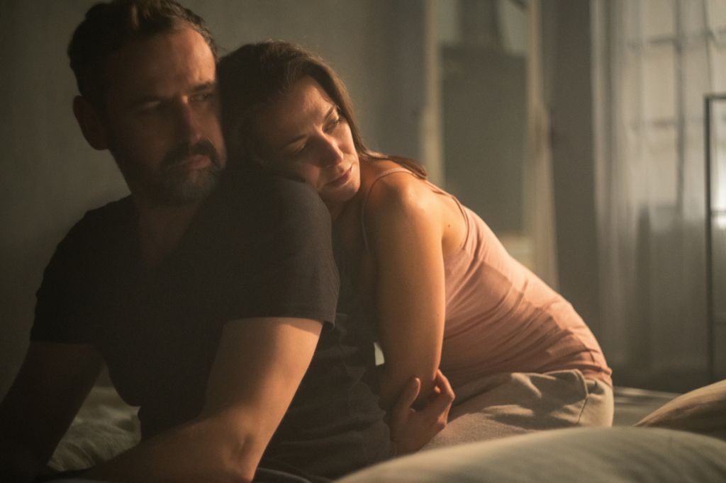 woman cuddles up to man from behind in bedroom, both looking into distance