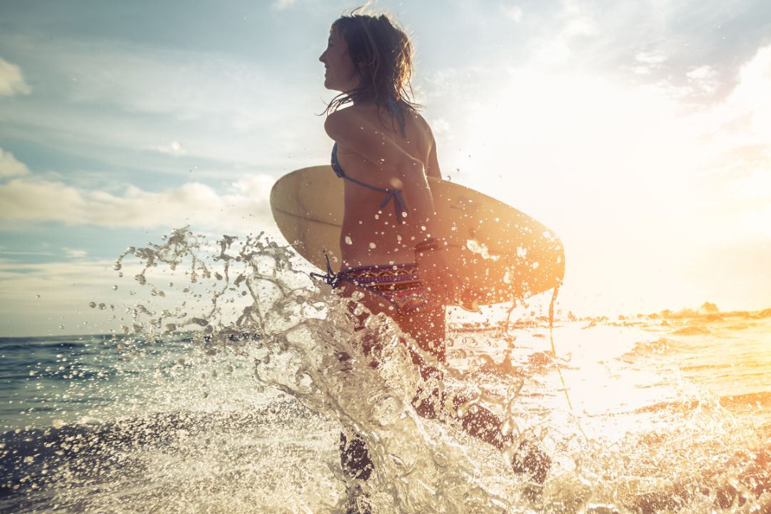 woman runs through the waves carrying a surfboard