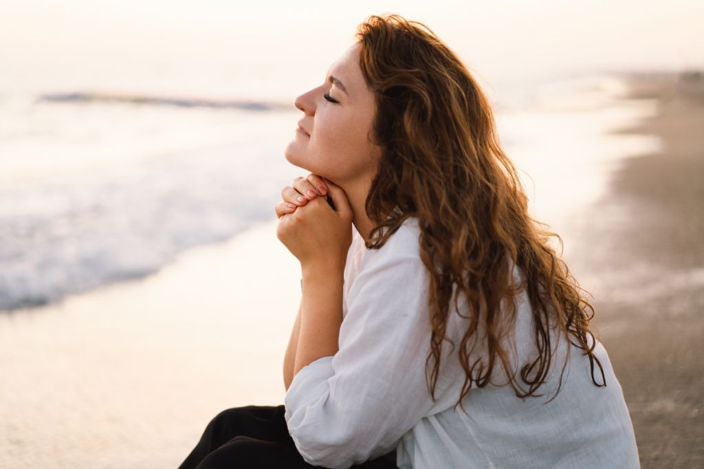 woman sits on the beach with her eyes closed and her hands under her chin in a prayer pose