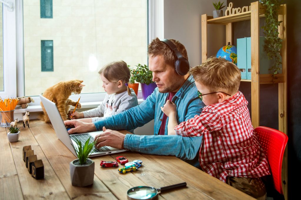 Dad looks stressed while managing two kids and working on laptop with headphones on. son tries to get his attention