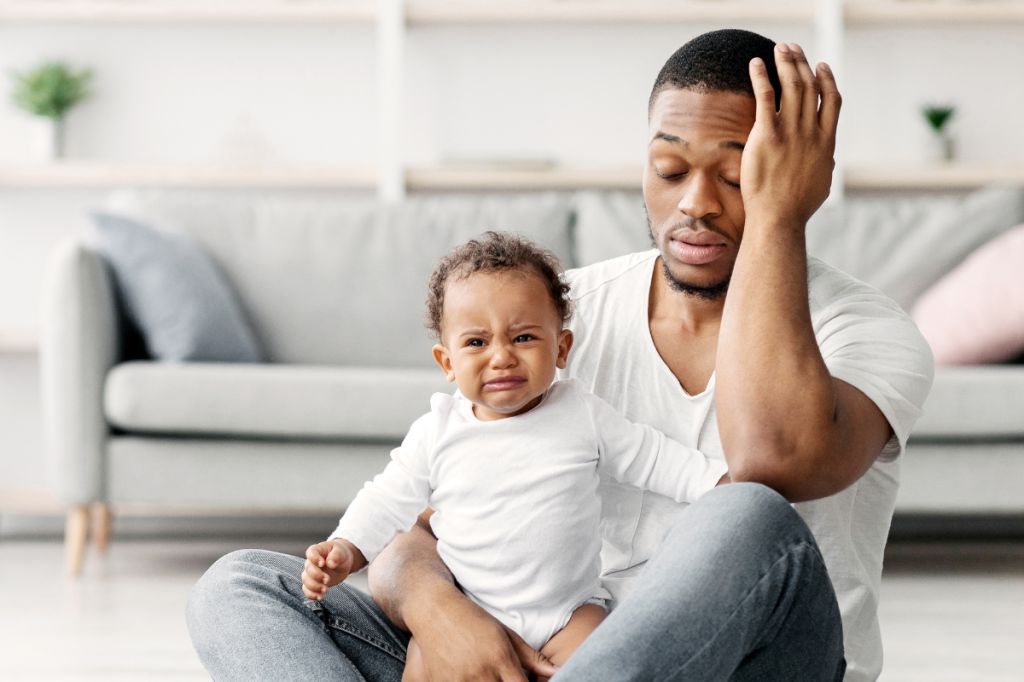 tired young dad sits on floor holding crying baby