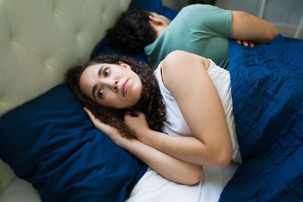 woman lies in bed looking into distance, her partner with his back to her. She is choking back tears of frustration and sadness