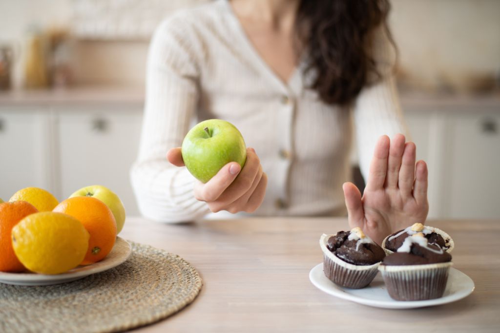 woman picks up green apple from plate of fruit, puts up hand to block plate of iced chocolate cakes next to it