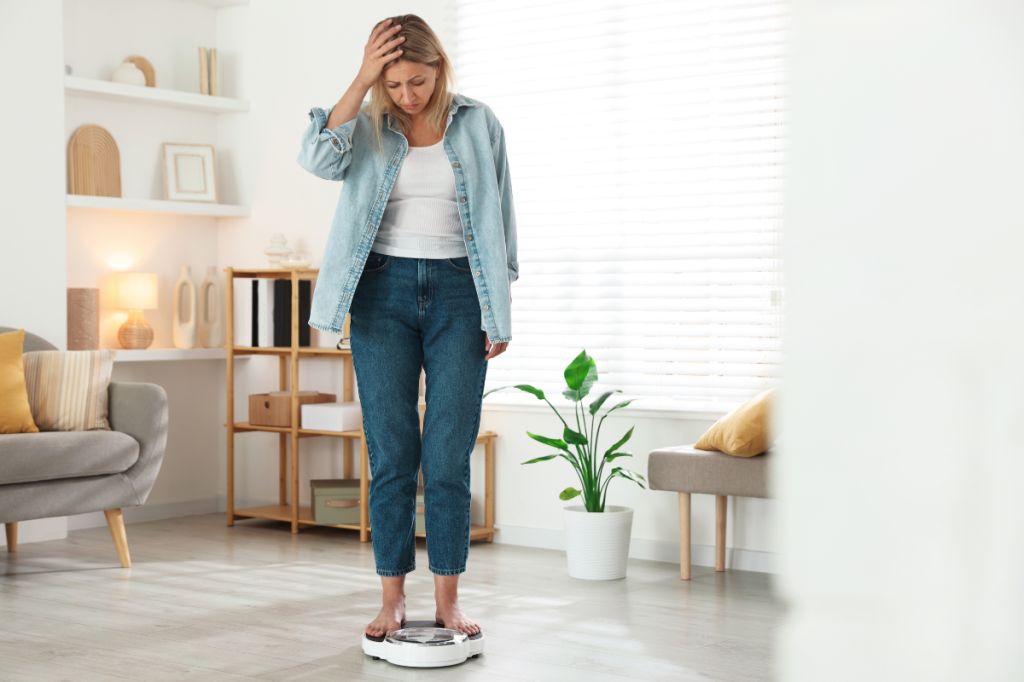 middle aged woman with blonde hair stands on scales in living room, hand on head, looking upset and disappointed at weight gain