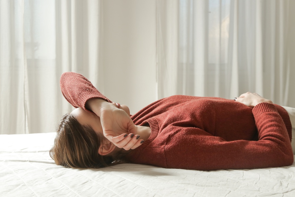 woman struggling with signs of burnout, she lays on a bed exhausted with arm covering face