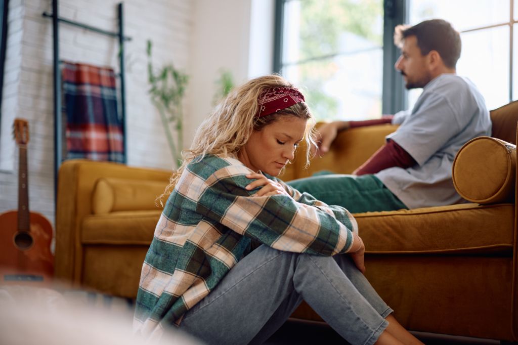 woman sits on floor by sofa. man looks into distance, she looks down at her arms. the silence is heavy with everything unsaid.