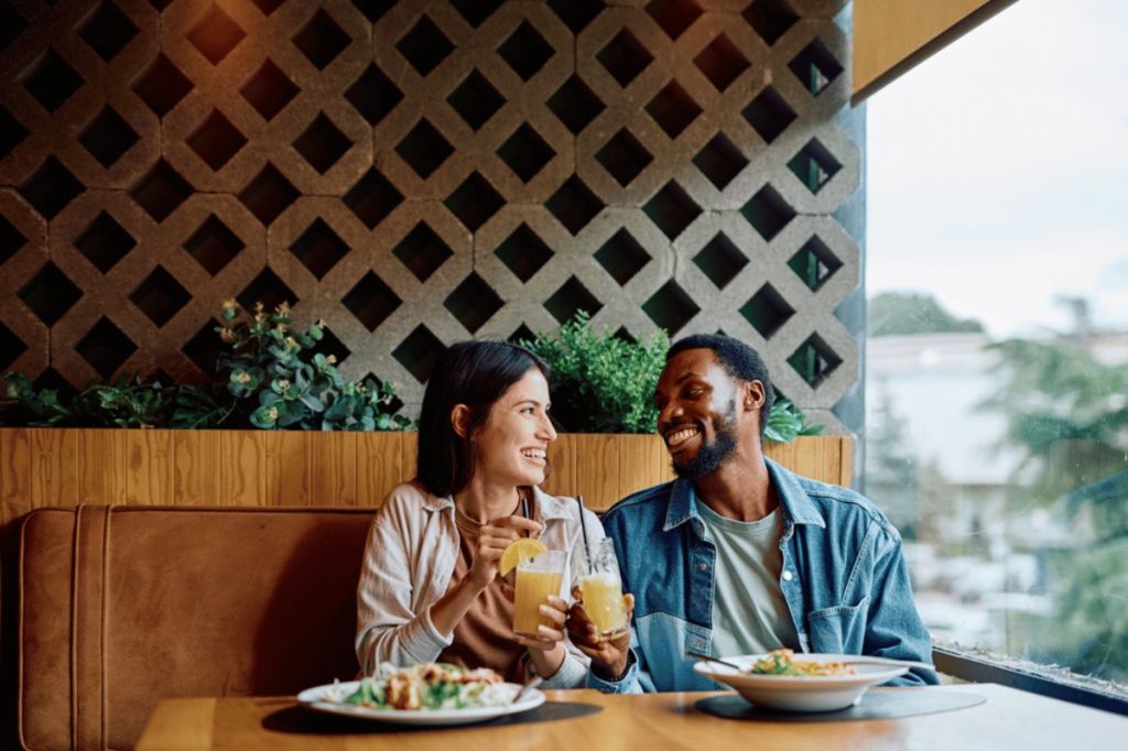 A man and woman sit next to each other in a restaurant. There are plates of food on the table. They hold their drinks to each other to say cheers and smile at one another.