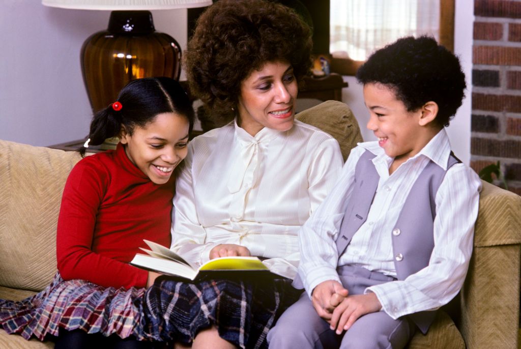 970s 1980s AFRICAN AMERICAN MOTHER SITTING ON COUCH WITH SON AND DAUGHTER READING TOGETHER