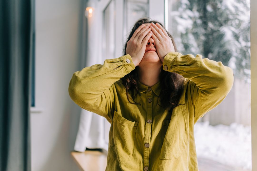 Woman covering her eyes with her hands by the window, expressing sadness and despair
