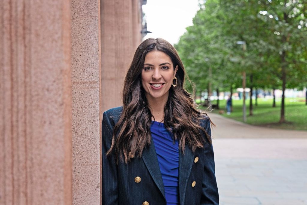 A white woman with long hair hair is smiling and leaning against a pink wall. There are trees in the background and she is wearing a blue top and navy blazer.
