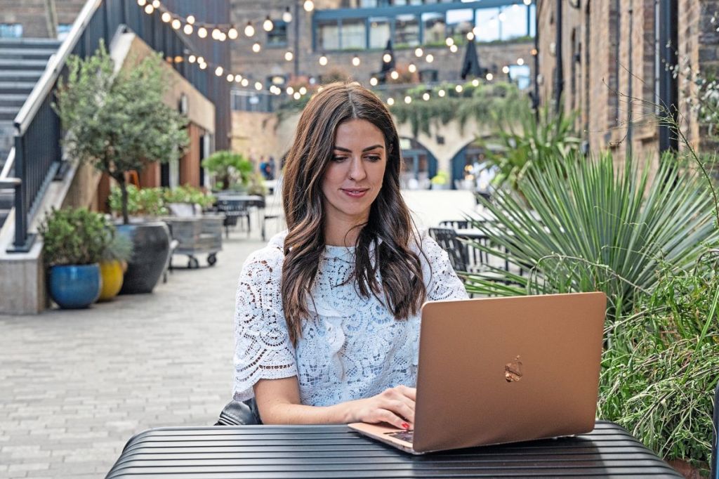 A white woman with long dark hair is sat outside at a table. She is wearing a white lacy top and typing on a laptop.