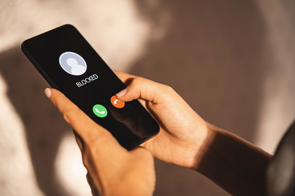  someone holding a phone -- the screen says 'BLOCKED NUMBER] is calling' and the person is pressing 'reject'