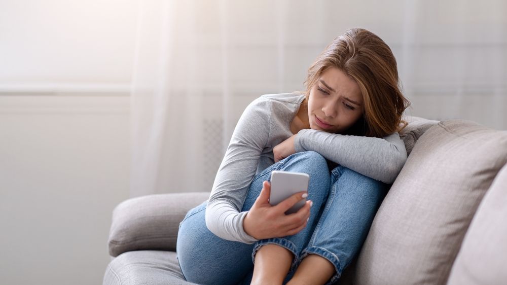 sad young woman looking at phone, curled up on sofa