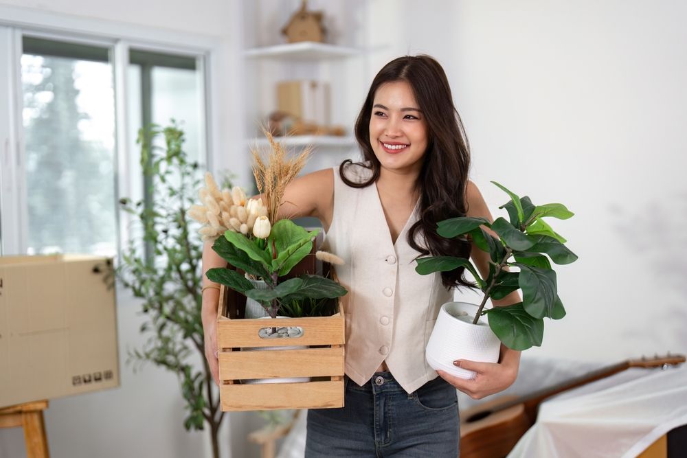 smiling woman holding boxes of plants in a new apartment