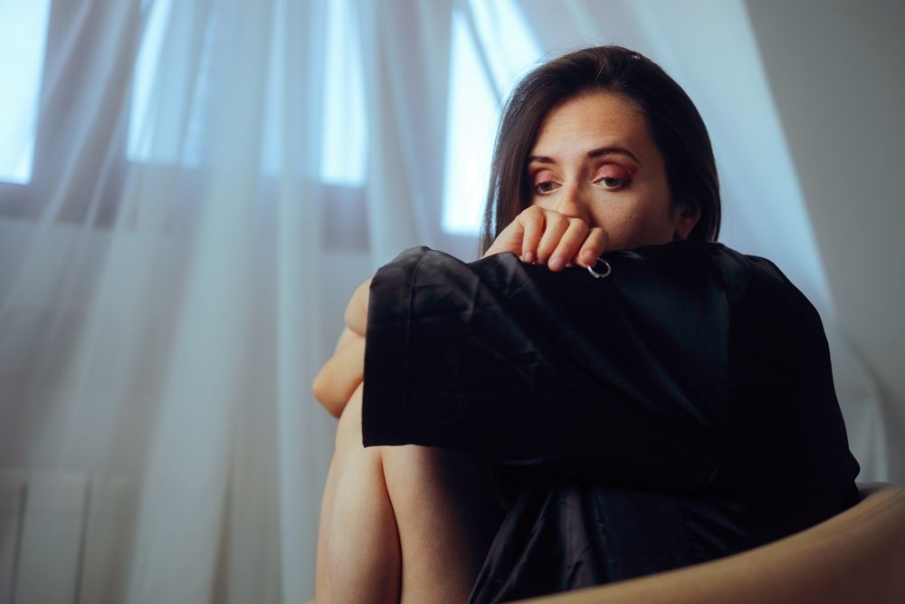 Woman looking unhappy in front of a window, knees pulled to her chest