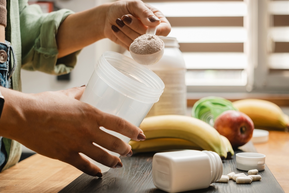 an image of hands with painted nails, putting powder into a smoothie, with various fruits in the background