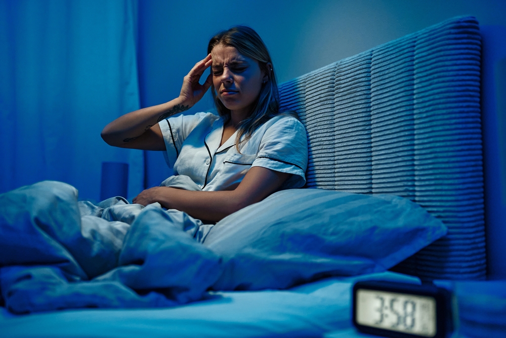 A woman in pyjamas in bed, clutching her head at night and looking unhappily at the clock