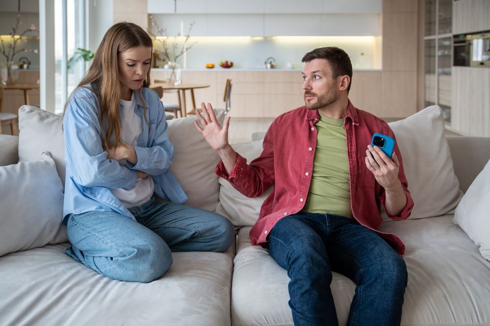 woman kneeling on the sofa with crossed arms, upset at man who is holding a phone and looking defensive