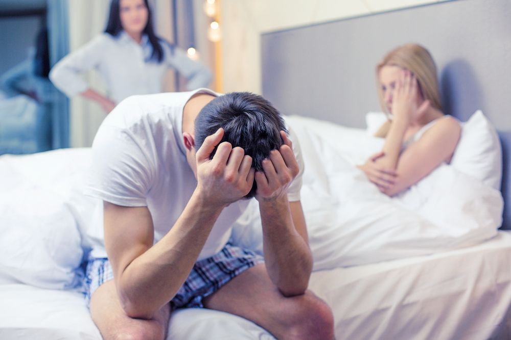 a woman stands in the doorway of a bedroom, a man looks distressed in bed with another woman